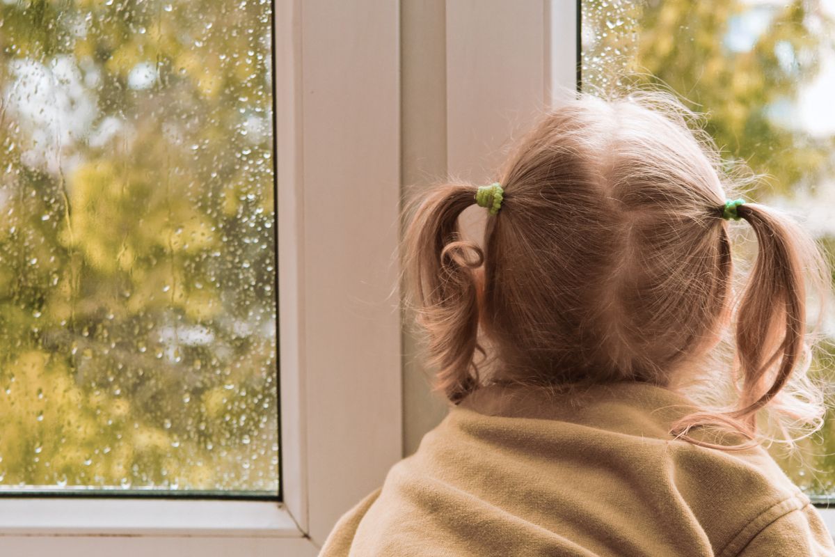 Kind sitzt am Fenster und schaut durch eine verregnete Fensterscheibe nach draußen